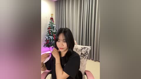 Media: Video of an East Asian woman with short black hair in a black top, sitting in a pink chair, looking at a decorated Christmas tree with red and green ornaments. Background features gray curtains and a white and black patterned pillow.