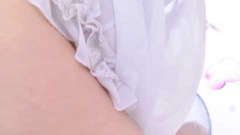 Media: A close-up video of a person's upper thigh and hip, showing a white, sheer, lace-trimmed garment partially covering the skin. The background is blurred, with soft pink hues.