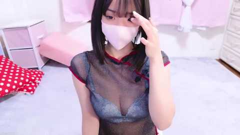 Media: Video of an Asian woman with short black hair, wearing a sheer black lace dress with red trim, a white mask, and a white face shield, in a pink-themed bedroom.