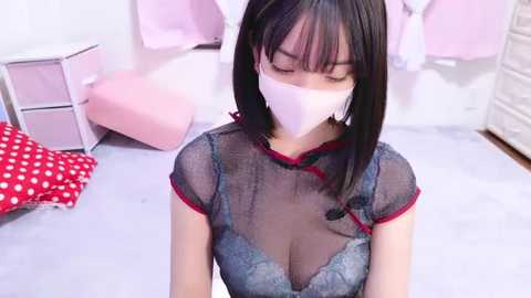 Media: Video of an Asian woman with straight black hair and a white face mask, wearing a sheer black top revealing a lace bra, standing in a pastel-colored room with pink pillows and polka-dotted furniture.