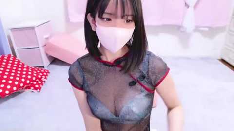 Media: Video of an Asian woman with straight black hair, wearing a sheer black dress revealing her large breasts, and a white face mask, in a pastel-pink room with a bed and red polka-dotted pillow.