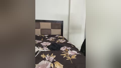 Media: Video of a simple bedroom with a bed featuring a dark brown quilt with large pink and yellow floral patterns. The headboard is a dark brown padded square. A black cat is lying on the bed, and a white object, possibly a shoe, is partially visible.