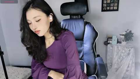 Media: Video of an Asian woman with long black hair, wearing a purple blouse, sitting in a black gaming chair, eyes closed, in a minimalist room with grey walls, framed art, and a small table with a plant.