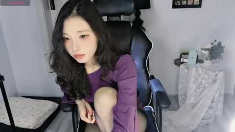 Media: A video of an Asian woman with long black hair, wearing a purple dress, sitting on a gaming chair in a minimalist room with white walls and a white rug.