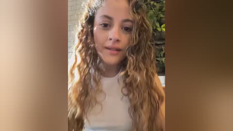 Media: A candid video of a young woman with long, curly blonde hair, wearing a white tank top, standing indoors with a blurred brick wall and green foliage in the background.