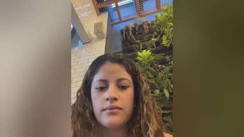 Media: Video of a young woman with curly brown hair, light brown skin, and neutral expression, standing in a modern indoor setting with white brick walls, a green wall of plants, and a wooden-framed window.