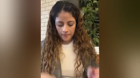 Media: Video of a young woman with curly, wavy brown hair, wearing a white top, sitting indoors next to a green plant and a white-tiled wall. She appears focused on her laptop.