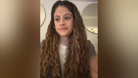 Media: A video of a young woman with long, curly, light brown hair, wearing a white shirt and dark jacket, standing indoors with white walls and round mirrors in the background.