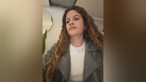 Media: A video of a young woman with curly, light brown hair, wearing a gray trench coat over a white turtleneck. She stands indoors, with a blurred background showing a plant and a round mirror.