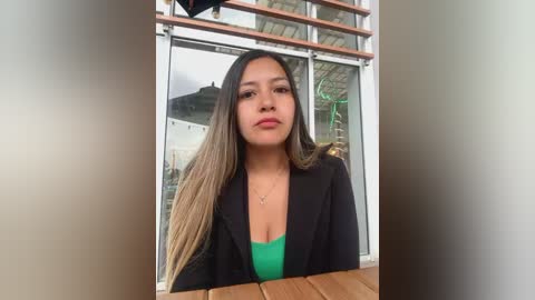 Media: Video of a Latina woman with long, straight, light brown hair and a green top, sitting indoors. She has a medium complexion and wears a black blazer. The background features a glass window with a blurred view of an outdoor scene.