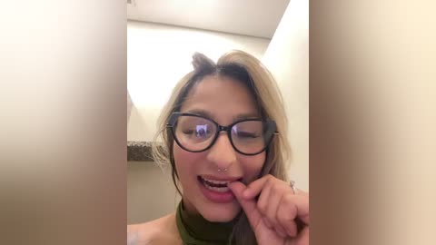 Media: Video of a young Asian woman with light brown hair, wearing black-framed glasses, a nose ring, and a green turtleneck, playfully biting her finger while smiling.