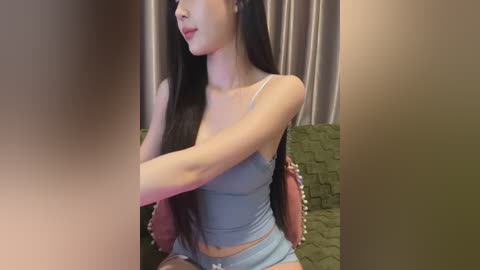 Media: Video of an Asian woman with long black hair, wearing a light blue spaghetti-strap tank top and matching shorts, seated on a green couch, partially obscured by a blurred figure in the foreground.