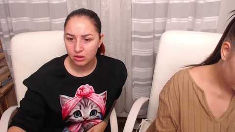 Media: Video of two women seated in a room with light-colored curtains and white chairs. One wears a black T-shirt with a pink cat design, while the other wears a beige top.