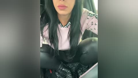 Media: Video of a woman with long black hair, wearing a pink sweater with black patterns, and black leather pants, sitting on a black car seat, with blurred background.