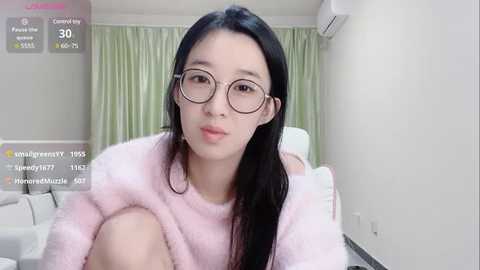 Media: Video of a young East Asian woman with long black hair, wearing round glasses and a pink fuzzy sweater, sitting on a white couch in a modern, minimally decorated room with green curtains.