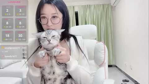 Media: Video of an East Asian woman with glasses, long black hair, and a gray cat, sitting in a white leather chair in a modern, bright room with light green curtains and a digital calendar.