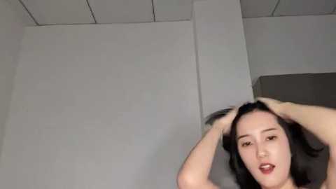 Media: Video of an Asian woman with straight black hair, light skin, and a neutral expression, hands in her hair, standing in a minimalist, modern room with white walls and a gray ceiling.