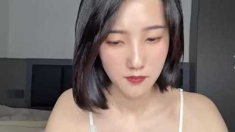 Media: Video of an East Asian woman with shoulder-length black hair, fair skin, wearing a white spaghetti-strap top, gazing downward. Background shows a bedroom with a bed and gray walls.