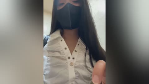 Media: A video of a woman with long dark hair, wearing a black face mask, white blouse, and black jacket, standing in an indoor, blurred background.