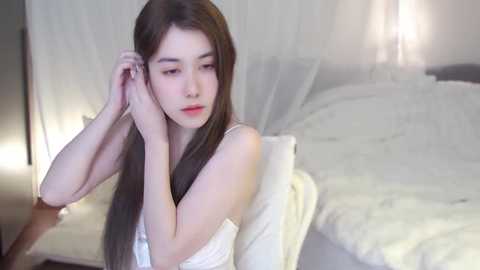 Media: A video of a young Asian woman with long, straight black hair, wearing a white tank top, sitting on a bed, adjusting her hair in a softly lit, minimalist bedroom.