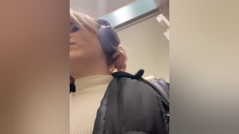 Media: Video of a woman with light skin, wearing a black puffer jacket and a white turtleneck, holding blue headphones, partially obscured by a blurry background in a sterile setting.