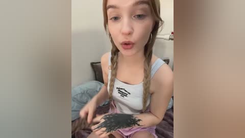 Media: Video of a young, fair-skinned girl with long blonde braids, wearing a white tank top with black text and pink shorts. She sits on a bed, drawing a black tattoo on her arm.