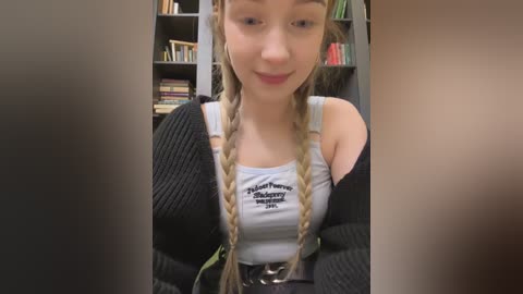 Media: Video of a young Caucasian woman with fair skin, wearing braided hair and a black sweater over a white tank top. She stands in a book-filled room with a casual, friendly expression.