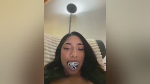 Media: Video of a young woman with long dark hair, eyes closed, gagged with a wad of money, lying on a bed with white and grey striped pillows, under a bright ceiling light.