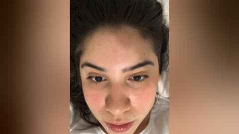 Media: Close-up video of a woman with light brown skin, dark brown hair, and thick eyebrows, lying on a bed. The background is blurred, focusing attention on her face.