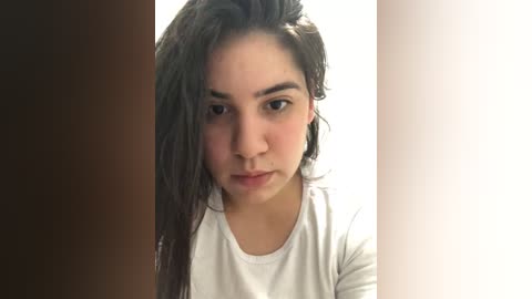 Media: Video of a young Latina woman with long dark hair, wearing a white shirt, standing in front of a plain white background. Her expression is neutral.