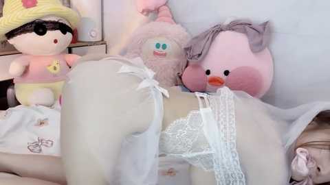 Media: Video of a person wearing white lace lingerie, lying face down on a bed, surrounded by plush toys, including a pink pig and a yellow octopus, in a softly lit, pastel-colored room.