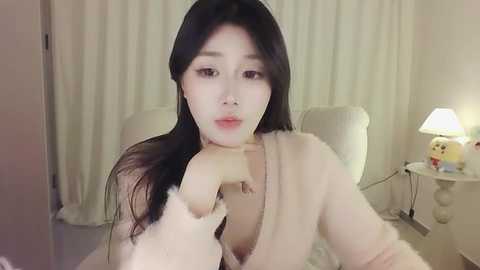 Media: Video of a young Asian woman with fair skin, long black hair, and a slight pout, wearing a cream-colored fluffy robe, posing in a softly lit, minimalist bedroom with white curtains and a nightstand lamp.