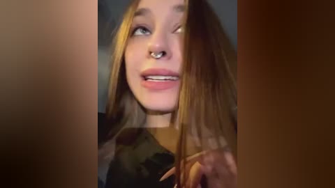 Media: A close-up video of a young woman with long, straight blonde hair, fair skin, and a septum piercing. She wears a black top and has a playful, slightly mischievous expression. The background is dark, focusing attention on her face.