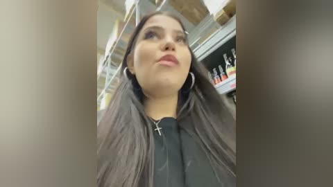 Media: A video of a young woman with long, straight, dark hair, wearing a black jacket and a cross necklace, looking upward in a grocery store aisle with shelves filled with products.