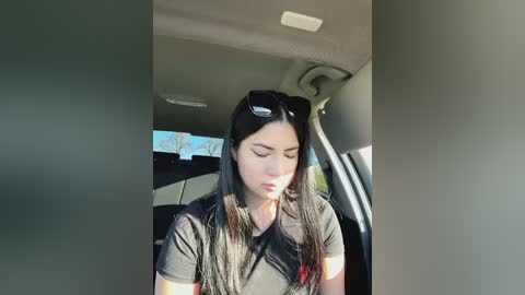 Media: Video of a young woman with long dark hair, wearing a black t-shirt and sunglasses, sitting in a car. She appears to be in a relaxed state, with her eyes closed and head tilted slightly to the right. The car interior is modern and well-lit.