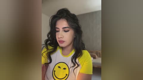 Media: Video of a young woman with long, wavy black hair, wearing a yellow T-shirt with a smiling yellow face graphic. She stands in a dimly lit room with a blurred background.