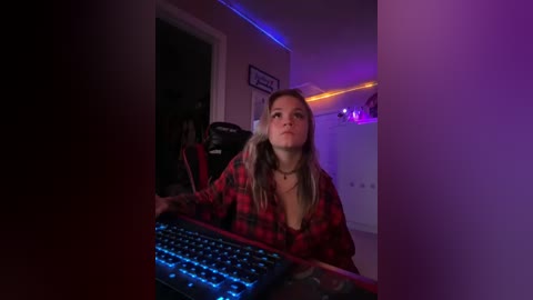 Media: Video of a young woman with long blonde hair, wearing a red plaid shirt, sitting at a desk with a blue-lit keyboard, in a dimly lit room with purple neon lights.