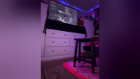 Media: Video of a dimly lit room with a flat-screen TV displaying a video, a black table, and a white dresser against purple LED strip lighting on the ceiling.