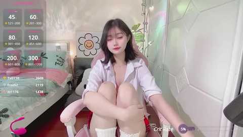 Media: A video of a young East Asian woman with fair skin, wearing a white shirt and thigh-high socks, sitting in a bedroom with a digital overlay displaying her stats and game information.