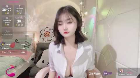 Media: A video of an East Asian woman with shoulder-length black hair, wearing a white shirt unbuttoned to reveal her breasts, sitting in a pastel-colored room with a bed and flower wall art.