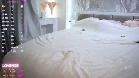 A video of a well-made bed with white linens in a modern, softly lit bedroom, featuring a white headboard and a wall with a floral mural. A white vanity table with a lamp is seen in the background.