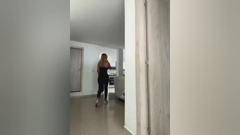 Media: A video of a blonde woman in a black tank top and pants walking away from the camera down a minimalist hallway with light wood floors and white walls.