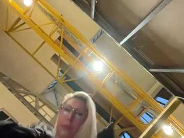 Media: Video of a blonde woman with glasses, wearing a black jacket, standing in an industrial building with a yellow metal walkway and exposed beams, fluorescent lights illuminating the scene.