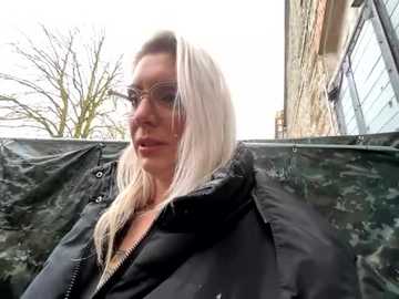 Media: A video of a blonde woman in a black puffy jacket, wearing reflective sunglasses, standing outside next to a brick building with a tree in the background.