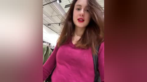 Media: Video of a young woman with long brown hair and fair skin, wearing a pink sweater, standing in a modern, brightly lit airport terminal.