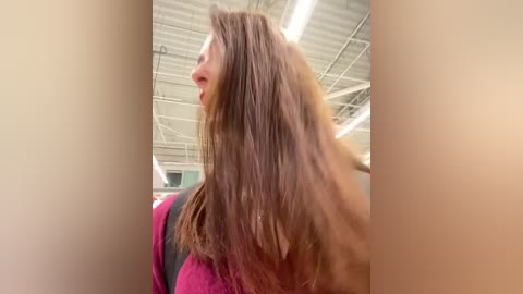 Media: A video of a young woman with shoulder-length brown hair, wearing a magenta sweater, taken indoors in a modern, industrial space with exposed ceiling beams and fluorescent lights.