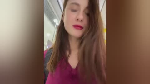 Media: A video of a young woman with long brown hair and fair skin, wearing bright red lipstick and a pink blouse, captured in a blurred, indoor setting.