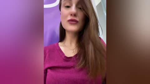 Media: Video of a young Caucasian woman with fair skin, straight brown hair, wearing a magenta sweater. Background features purple and beige tones. She has a neutral expression and is slightly out of focus.