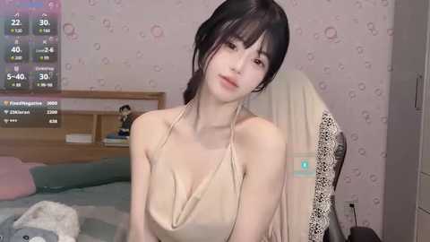 Media: A video of a slender East Asian woman with fair skin and dark hair, wearing a beige halter top, sitting in a bedroom with a pink, patterned wallpaper, a wooden bed, and a digital clock.