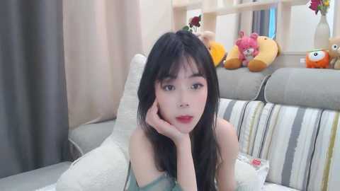 Media: Video of an Asian woman with long black hair, resting her chin on her hand, in a cozy living room with a beige sofa, stuffed animals, and light-colored curtains.